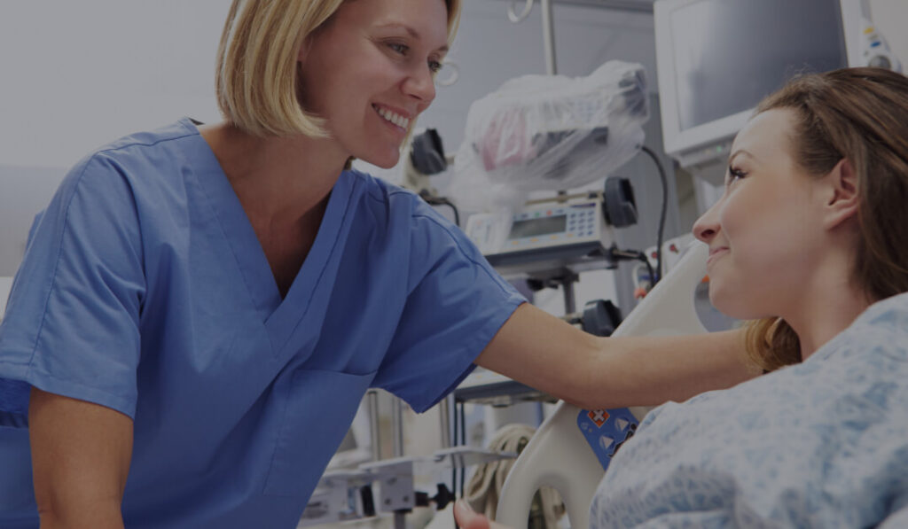 A smiling nurse in scrubs leans over a patient in a hospital bed in a facility representing Saint Luke’s Health System